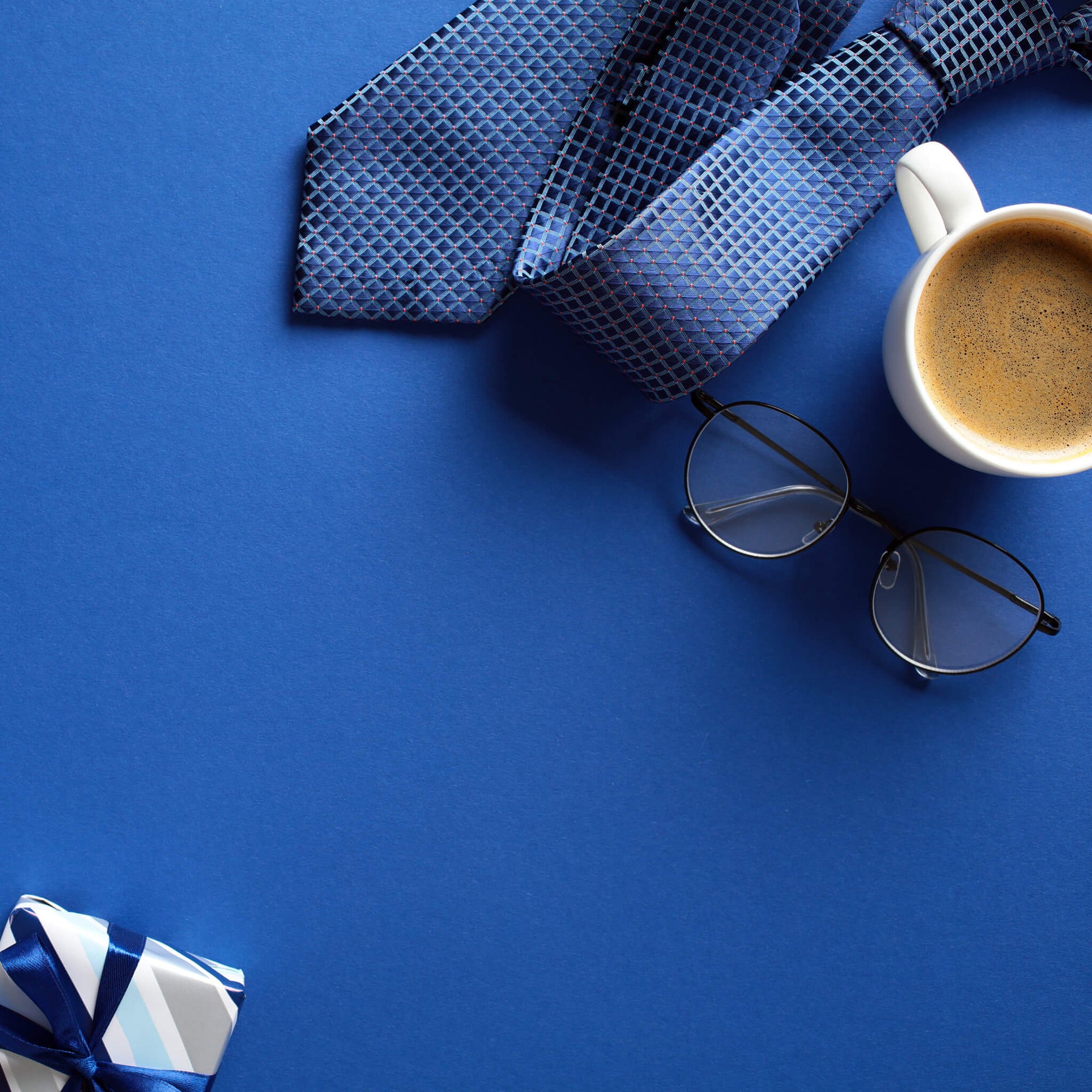 Flat lay of a tie, coffee cup, eyeglasses, and wrapped gift on a blue background—symbolizing stylish and functional gifts for business dads.