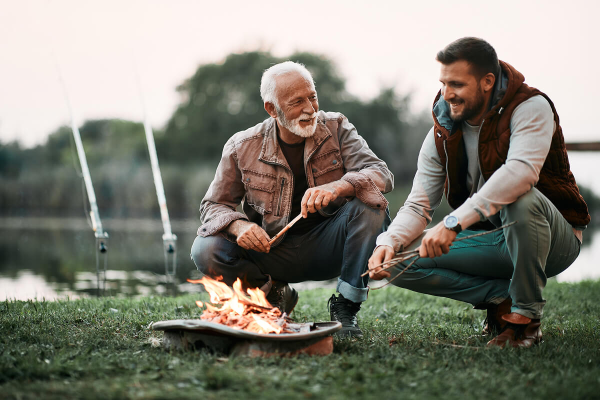 Dad standing by a fire pit holding a Bacon Bouquet and ammo can full of jerky, embodying the ultimate outdoor gift experience.