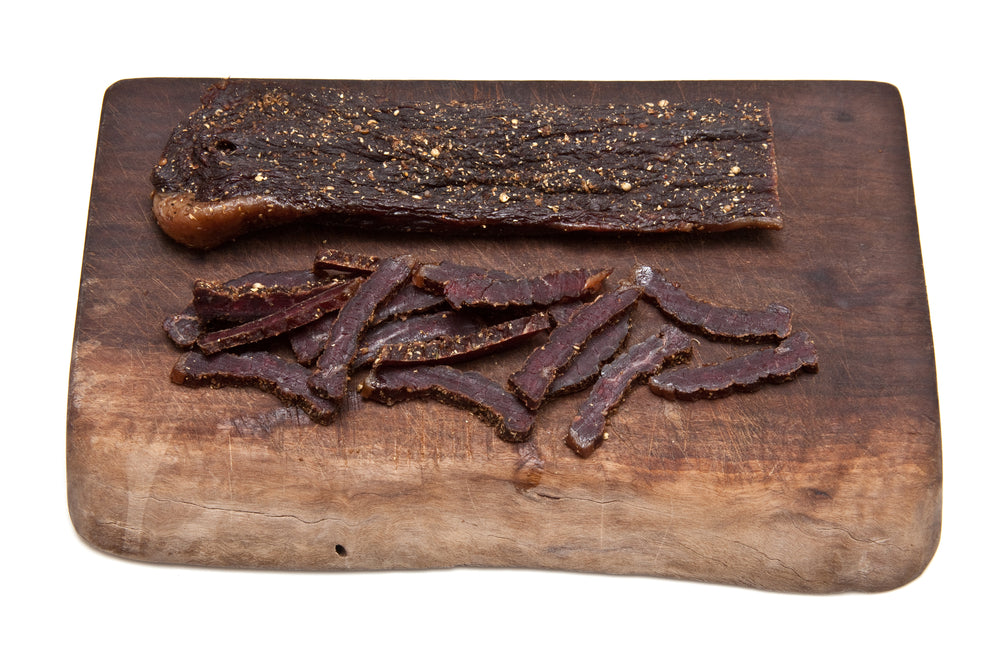 two pieces of biltong on a wood cutting board, one piece has been sliced and the other is whole