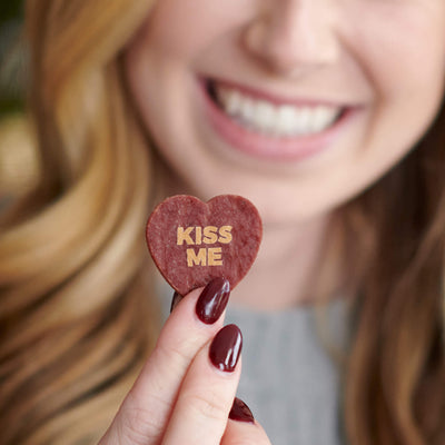 A smiling woman holding a heart-shaped piece of beef jerky with the words 'KISS ME' printed on it, showcasing a playful and edible Valentine's Day gift.