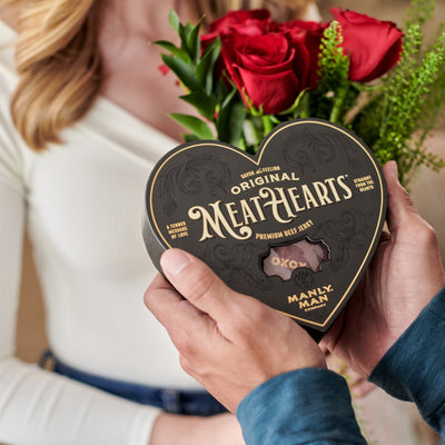 A person holding a black heart-shaped box of 'Original MeatHearts' beef jerky while presenting it to a woman holding a bouquet of red roses, creating a unique Valentine's Day gifting moment.