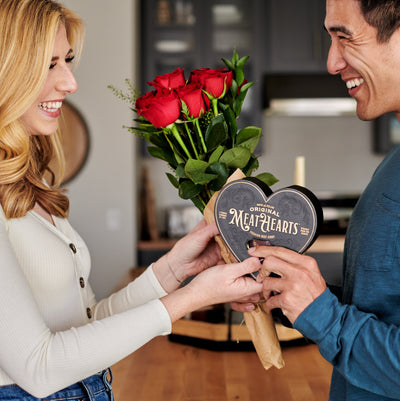 Alt text: "A smiling couple exchanging unique Valentine's Day gifts, with the man holding a box of 'Original MeatHearts' beef jerky in a heart-shaped package and the woman presenting a bouquet of red roses."