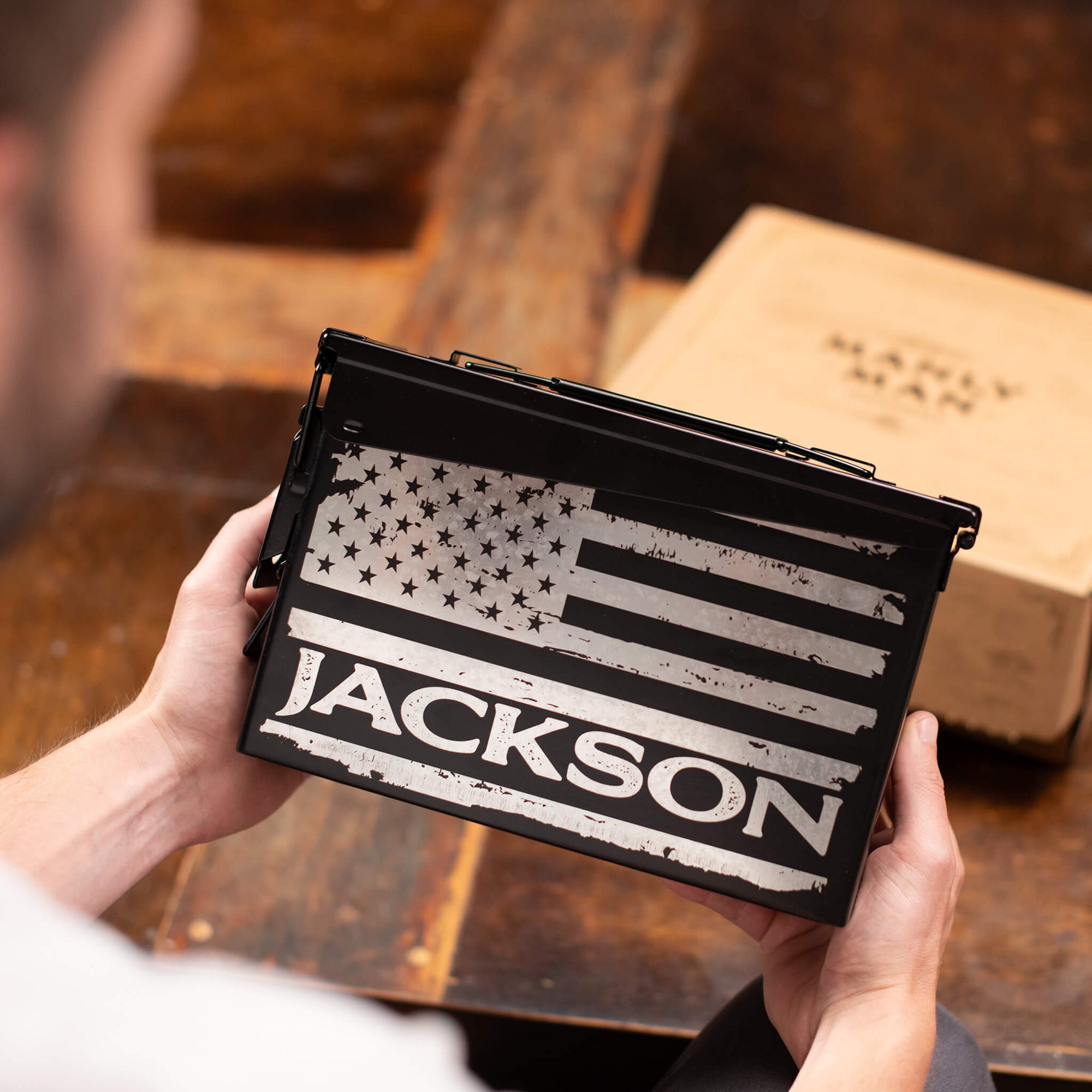 A person holding a black personalized ammo can with a distressed American flag design and the name "Jackson" printed on it. A Manly Man Co. gift box sits on a rustic wooden table in the background.