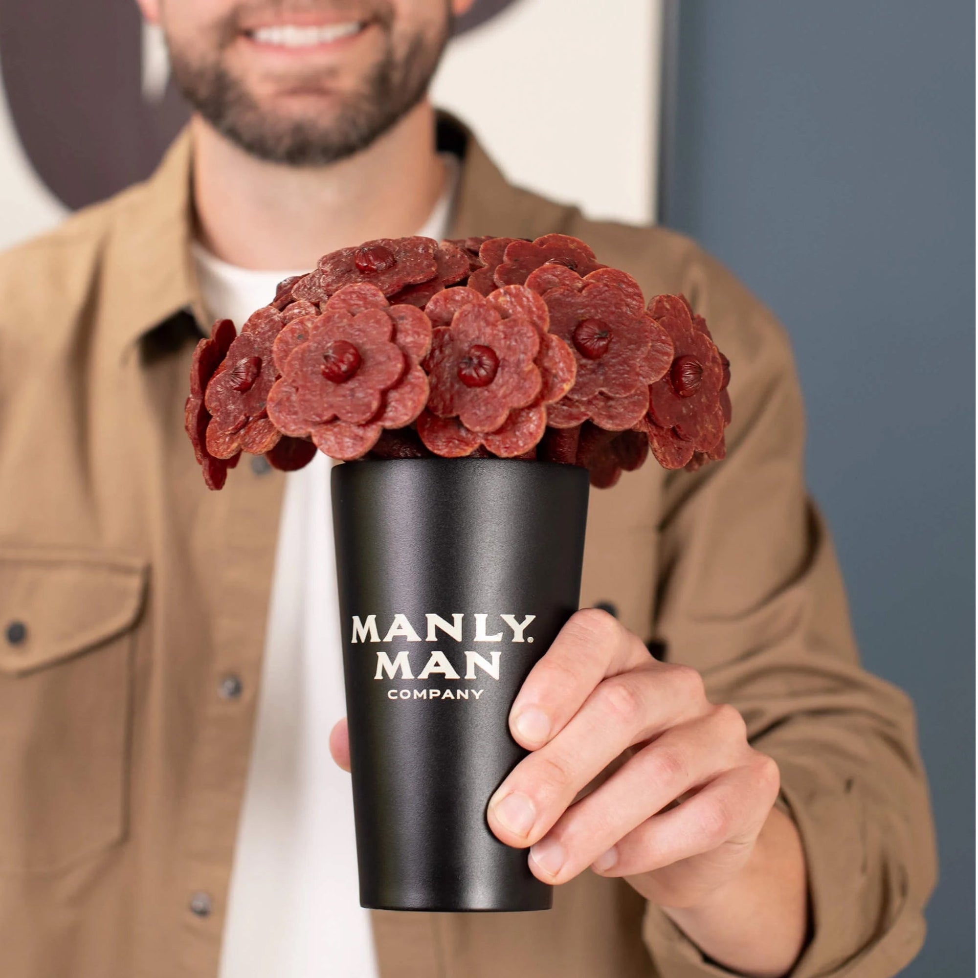 A man holding a black Manly Man Company pint glass filled with a bouquet of beef jerky shaped like flowers, offering a unique and masculine gift idea.