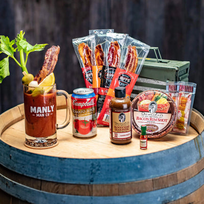 A Bloody Mary in a Manly Man Co. glass mug, garnished with bacon, celery, olives, and lemon, on a wooden barrel. Surrounding it are bacon jerky packs, tomato juice, Bloody Mary seasoning, bacon rim salt, a mini Tabasco bottle, and olives.