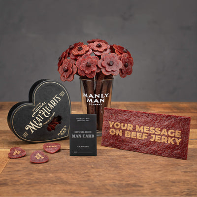 A bundle of Manly Man Company gifts featuring a beef jerky flower bouquet in a pint glass, a black 'Original MeatHearts' box, a personalized beef jerky card with a custom message, and an 'Official Issue Man Card,' displayed on a rustic wooden surface.