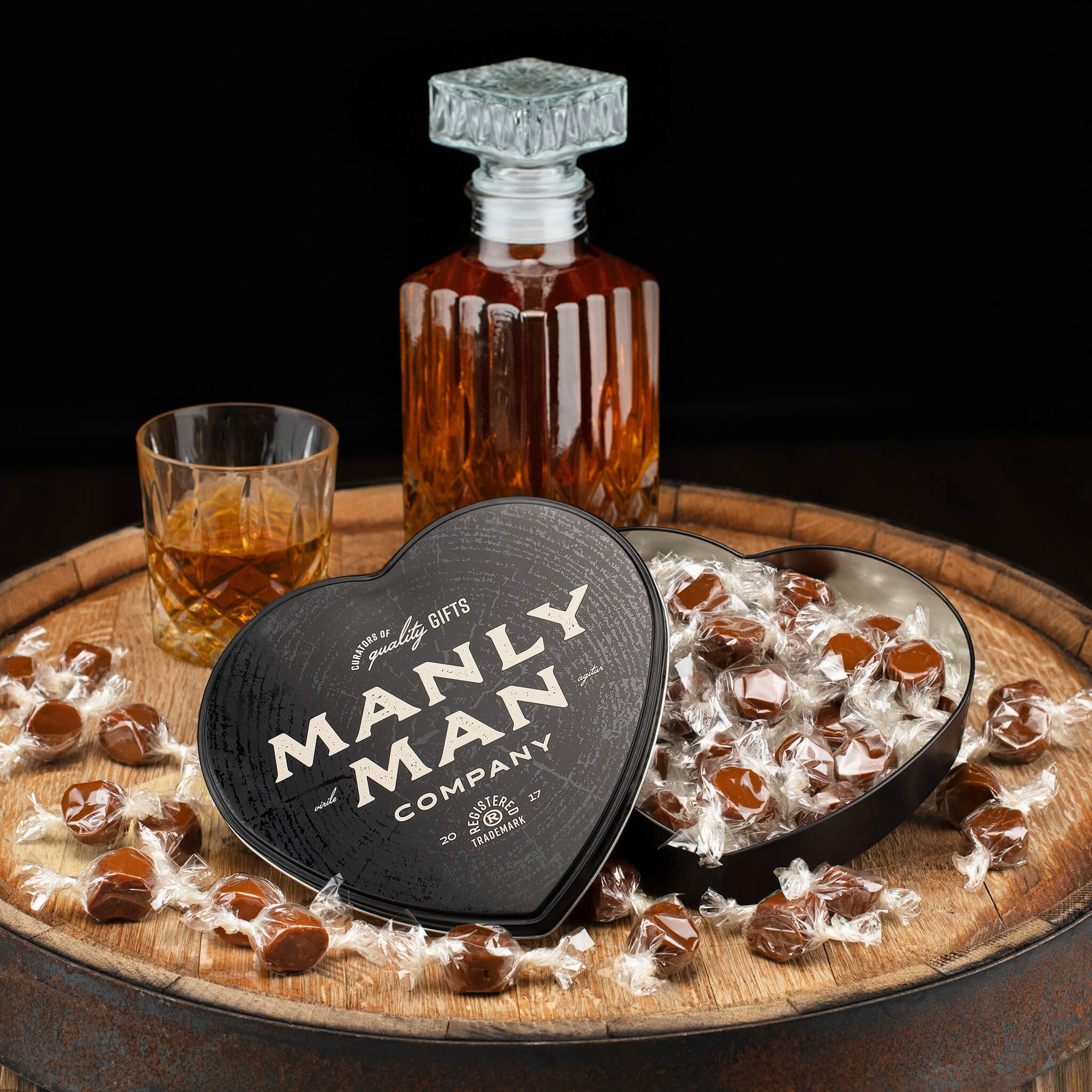 A black heart-shaped Manly Man Company tin filled with individually wrapped whiskey caramels, displayed on a rustic wooden barrel alongside a glass decanter and a tumbler of whiskey.