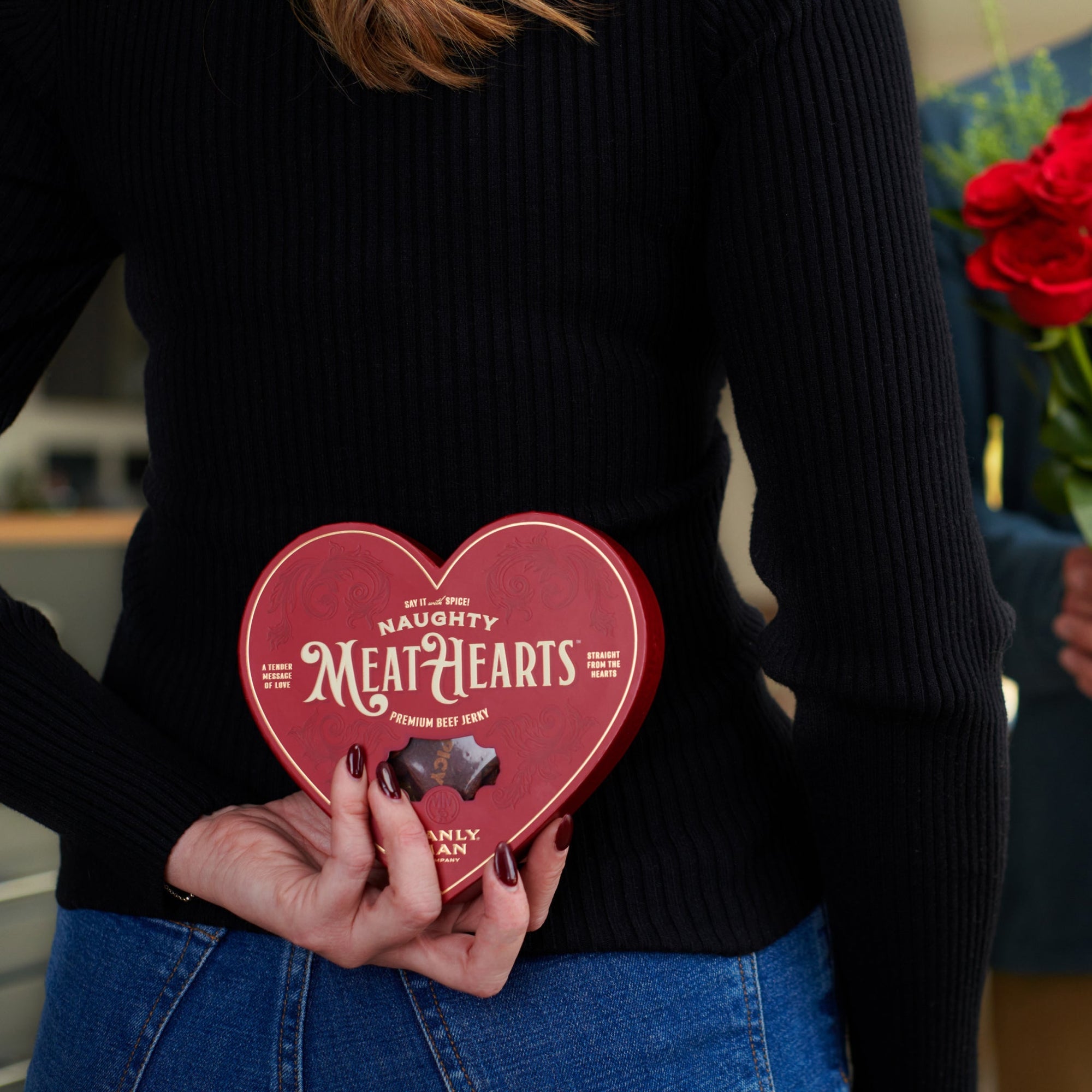 A woman in a black sweater holding a red heart-shaped box of 'Naughty MeatHearts' beef jerky behind her back while a man in the background holds a bouquet of red roses