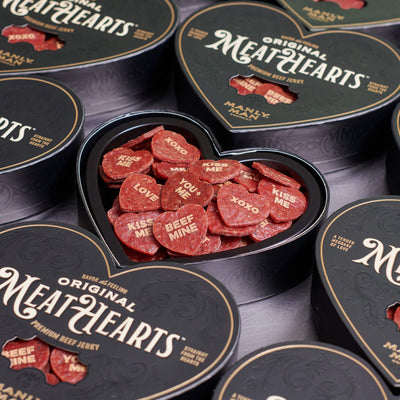 A collection of 'Original MeatHearts' heart-shaped black boxes filled with beef jerky pieces featuring playful messages like 'KISS ME,' 'XOXO,' 'LOVE,' and 'BEEF MINE,' perfect for a unique and savory Valentine's Day gift.