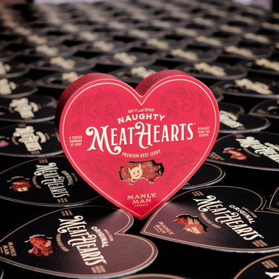 A vibrant red heart-shaped box of 'Naughty MeatHearts' premium beef jerky prominently displayed in front of a backdrop filled with black 'Original MeatHearts' boxes, showcasing playful and savory Valentine's Day gift options.