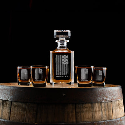 A personalized whiskey decanter set featuring an American flag design and custom engraving. Includes a decanter and four matching glasses, displayed on a rustic barrel with a dark background. A bold and patriotic gift for whiskey lovers.