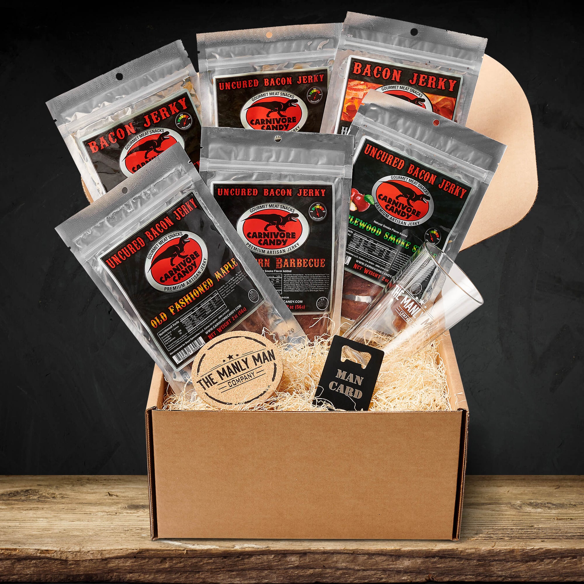 Gift box filled with bags of bacon jerky, a coaster, man card, and pint glass, on wood table
