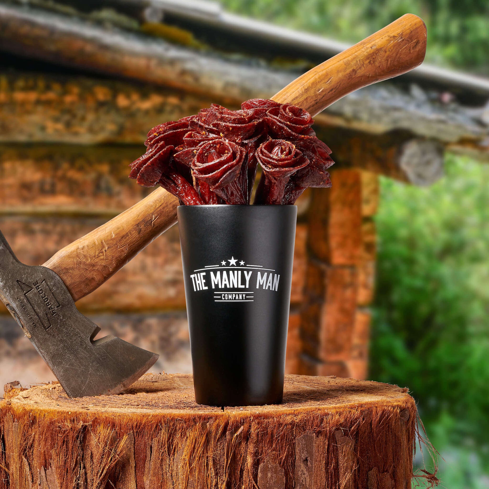 Beef jerky rose bouquet in Black Steel pint glass "vase" and hatchet on upright log, in front of cabin