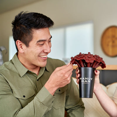 A smiling man in a green shirt reaching for a piece of beef jerky from a bouquet arranged in a black Manly Man Company pint glass, held by someone off-camera