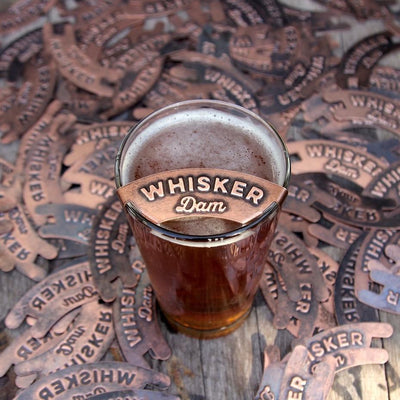 Whisker damn attached to glass, given as manly grooming gift, that is sitting on pile of copper whisker damns