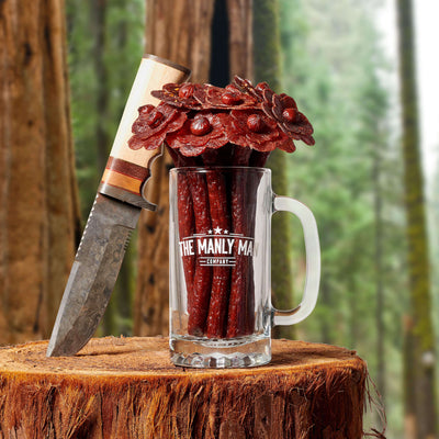 Beef jerky flower bouquet in mug pint glass "vase", next to Bowie knife, on log and forrest background